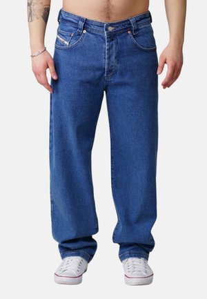 Mann in lockeren blauen Jeans und weißen Turnschuhen, steht mit entspannten Händen an den Seiten vor einem schlichten Hintergrund.