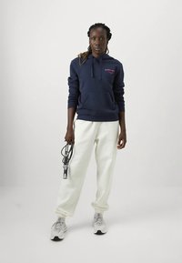 Γυναίκα που φοράει ναυτικό hoodie, λευκά παντελόνια φόρμας και λευκά αθλητικά παπούτσια κρατώντας ένα μαύρο και ασημί σχοινί αντίστασης μπροστά από ένα απλό ανοιχτό φόντο.