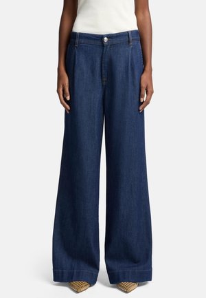 Persoon draagt donkerblauwe jeans met wijde pijpen en beige puntige schoenen, staat met ontspannen armen langs het lichaam.
