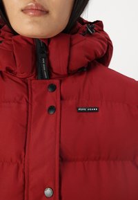 Crvena puffer jakna s visokim ovratnikom, crnim dugmadi na preklop i zatvaračem. Ima oznaku "Pepe Jeans London". Glatka tekstura.