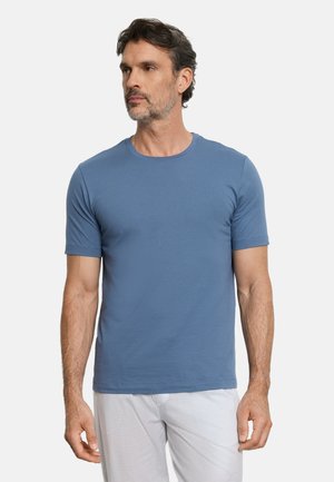 Mężczyzna z ciemnymi włosami i brodą, ubrany w prostą niebieską koszulkę z krótkim rękawem i jasne spodnie, zwrócony lekko w lewo.