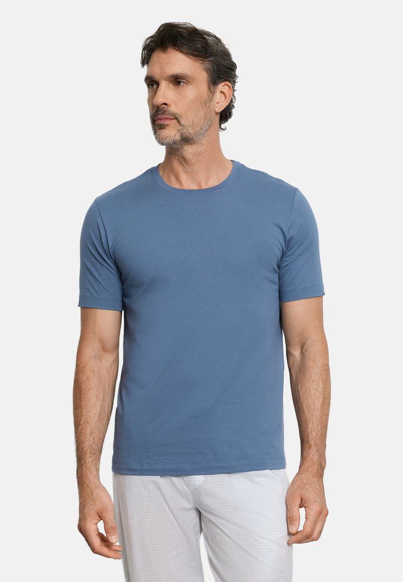 Homme aux cheveux foncés et barbe, portant un t-shirt bleu uni à manches courtes et un pantalon de couleur claire, légèrement tourné vers la gauche.