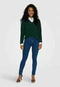 Swehter verde de malha sobre uma camisa branca, combinado com jeans justos azuis e sandálias pretas de salto. A modelo está em uma pose neutra.