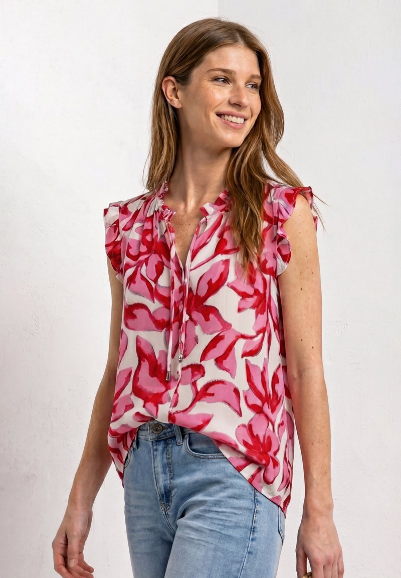 Jonge vrouw lacht, draagt een witte mouwloze blouse met rode bloemenprint en lichtblauwe spijkerbroek tegen een effen achtergrond.