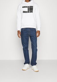 White hoodie with black "TOMMY JEANS" logo, paired with blue denim jeans. Footwear features white sneakers with gum sole.