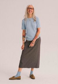 Světle modrý top s krátkým rukávem a kontrastními detaily, šedá sukně ke kotníkům, modré ponožky a hnědé semišové boty. Viditelné tetování na pažích.