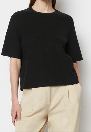 Vrouw draagt een zwarte, kort mouw gebreide trui gecombineerd met beige, hoog getailleerde broek, staand met één hand in de zak.