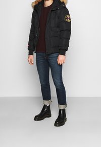 Czarna kurtka puchowa z kapturem obszytym futrem, suwakiem z przodu, dwoma kieszeniami na piersi i naszywką z logo. Noszona z ciemnymi dżinsami i czarnymi botkami za kostkę.