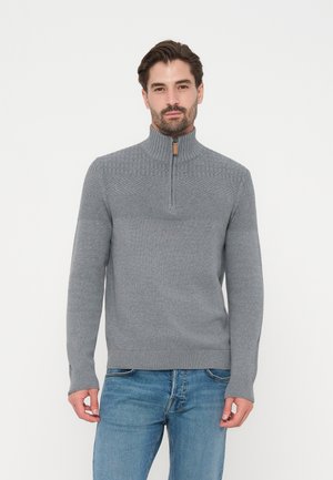 Mann trägt einen hellgrauen Strickpullover mit Viertel-Reißverschluss am Kragen und blaue Jeans, stehend vor weißem Hintergrund.