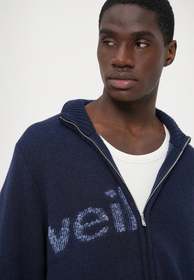 Marinblå ziptröja med ribbad krage; mjuk textur; har ljusblå "veil"-bokstäver tvärs över bröstet; lagd över en vit skjorta.