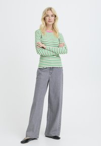 Frau mit langen blonden Haaren, die ein grün-weiß gestreiftes langärmliges Oberteil, graue weitgeschnittene Jeans und schwarze Schuhe trägt, steht mit verschränkten Armen.