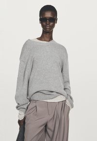 Grau gestrickter Pullover mit V-Ausschnitt, übergroßen Ärmeln, darüber ein helles, strukturiertes Oberteil; kombiniert mit hellgrauen, plissierten Hosen.