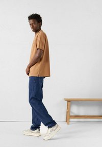 Polo marrone, jeans blu e sneakers leggere. La polo presenta maniche corte, spacchi laterali e una vestibilità comoda. Design semplice e materiali resistenti.