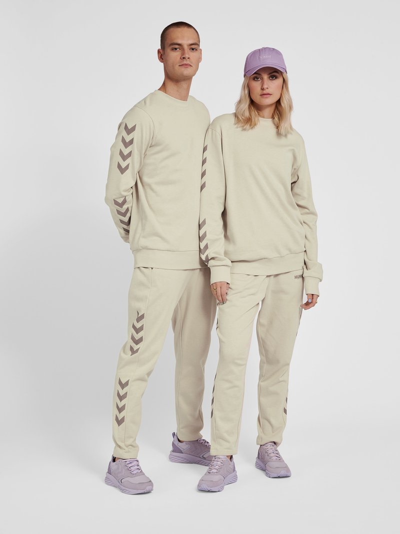 Zwei Personen tragen passende beige Trainingsanzüge mit Chevron-Mustern an Ärmeln und Hosen sowie hellviolette Sneakers und stehen nebeneinander.