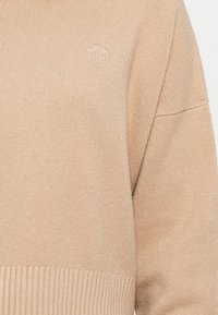 Pull en tricot beige avec un ourlet et des poignets côtelés. Présente un petit logo circulaire de couleur assortie à l'avant. Texture douce, design décontracté.