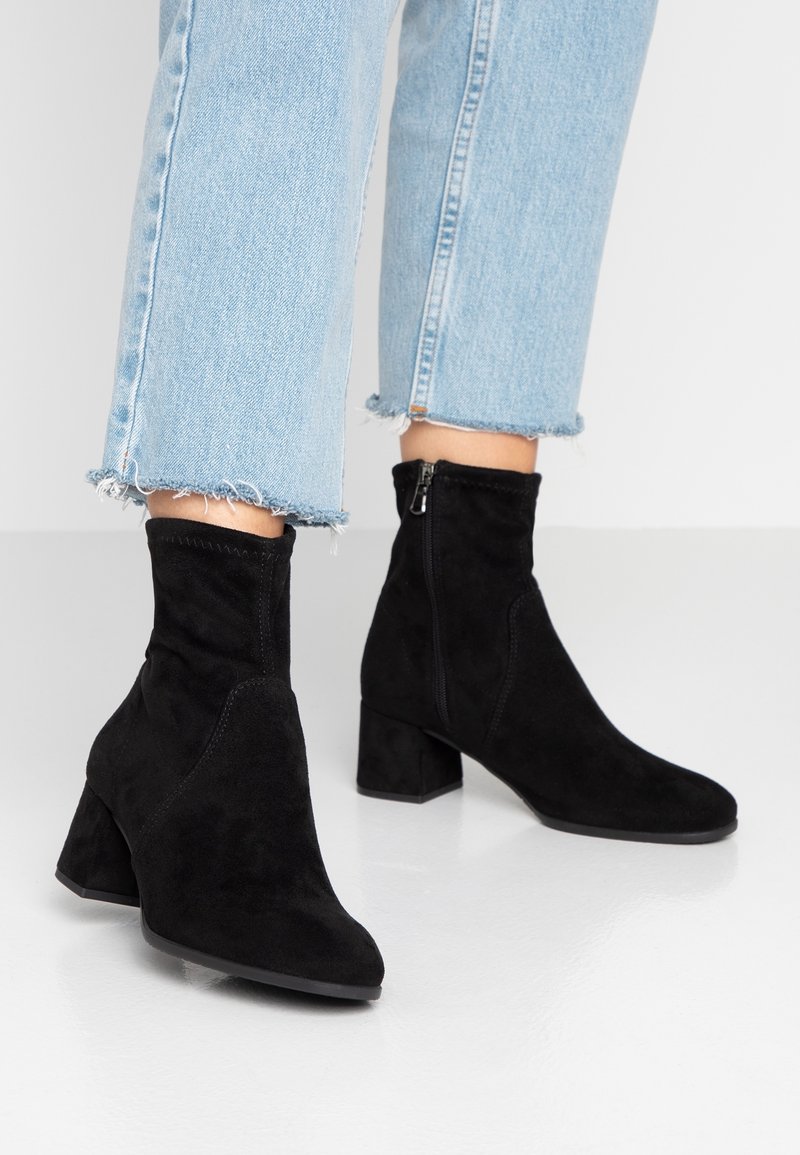 Bottines noires en daim avec un talon bloc, une fermeture éclair latérale et un bout pointu. Elles présentent une texture lisse et une silhouette structurée.