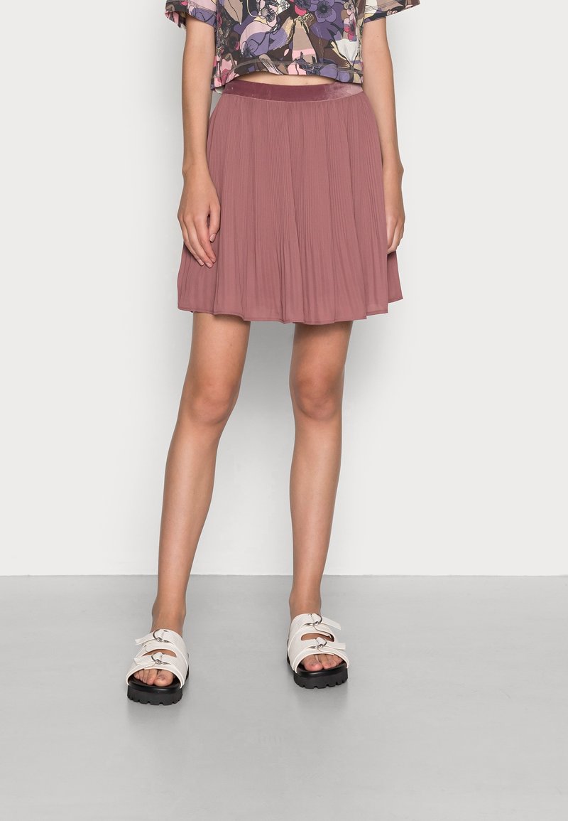Femme portant une jupe plissée mauve, un haut court à fleurs et des sandales blanches, debout sur un sol gris devant un mur blanc.