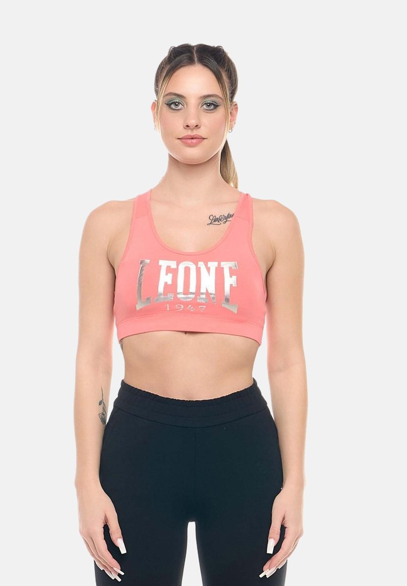 Jeune femme aux cheveux attachés porte un soutien-gorge de sport corail avec le texte « LEONE 1947 » et un legging d’entraînement noir, debout devant un fond uni.