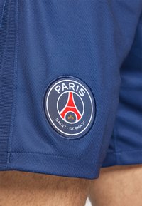 Marinsblå sportshorts med en cirkulär broderad logotyp med texten "PARIS" och "SAINT-GERMAIN", röda och vita detaljer samt en lysande lilja.