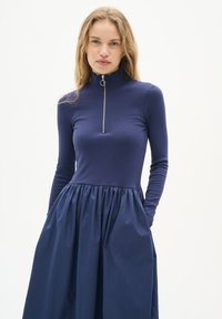 Rochie bleumarin cu un top fixat, din dantelă, și o fustă evazată, plisată; prezintă un guler înalt și un fermoar pe jumătate. Materialul pare neted și texturat.