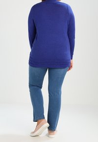 Femme portant un haut bleu à manches longues avec un ourlet texturé, un jean bleu et des chaussures beiges à enfiler, debout devant un fond clair uni.