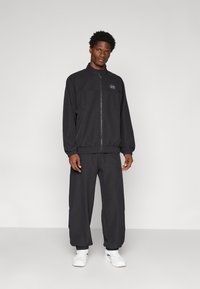 Un survêtement noir avec une veste zippée à col haut et des poignets élastiques, associé à un pantalon ample. Le matériau semble lisse et léger.