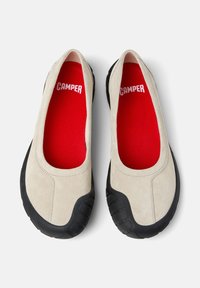 Chaussures slip-on en suède beige avec des semelles en caoutchouc noir. Les semelles intérieures rouges portent le logo "CAMPER". Texture lisse avec un design de bout arrondi.