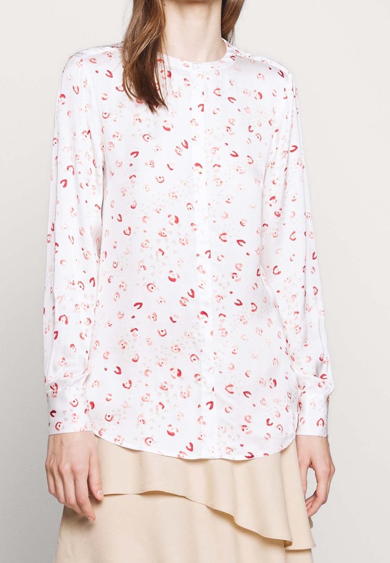 Blouse blanche à manches longues avec une texture lisse, ornée de motifs floraux rouges et abstraits, détails de boutons et ourlet arrondi.