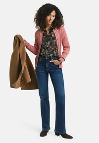 Rosa kabelgestrickte Strickjacke über einer blumigen Bluse, kombiniert mit blauen Schlagjeans und dunklen Wildleder-Ankle-Boots. Hält einen braunen Mantel.