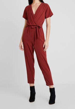 Combinaison couleur rouille avec un design portefeuille, un col en V plongeant, des manches courtes et une ceinture à nouer à la taille. Associée à des bottines noires à la cheville.