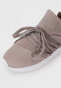 Hellgraue Sportschuh mit einer strukturierten Mesh-Oberfläche, auffälligem Schnürsystem und einer weißen Gummisohle mit subtiler Branding.