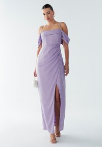 Femme portant une robe de soirée lavande à épaules dénudées avec une fente à la cuisse, tenant un petit sac à main blanc et portant des talons à brides transparentes.