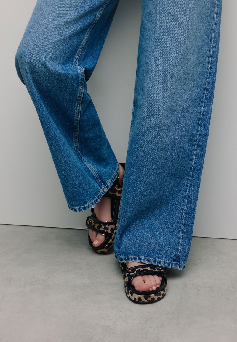Wijd-uitlopende lichte spijkerbroek met rafelige zoom, gecombineerd met zwarte sandalen met een panterprint en een chunky gespsluiting.