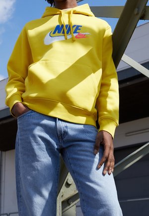 Personne portant un sweat à capuche Nike jaune vif et un jean bleu clair, debout à l'extérieur près d'une structure métallique sous un ciel dégagé.