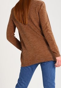 Brązowy, teksturowany sweter z dzianiny z długimi rękawami i luźnym krojem, zestawiony z niebieskimi dżinsami. Sweter ma subtelny wzór i prążkowany dół.