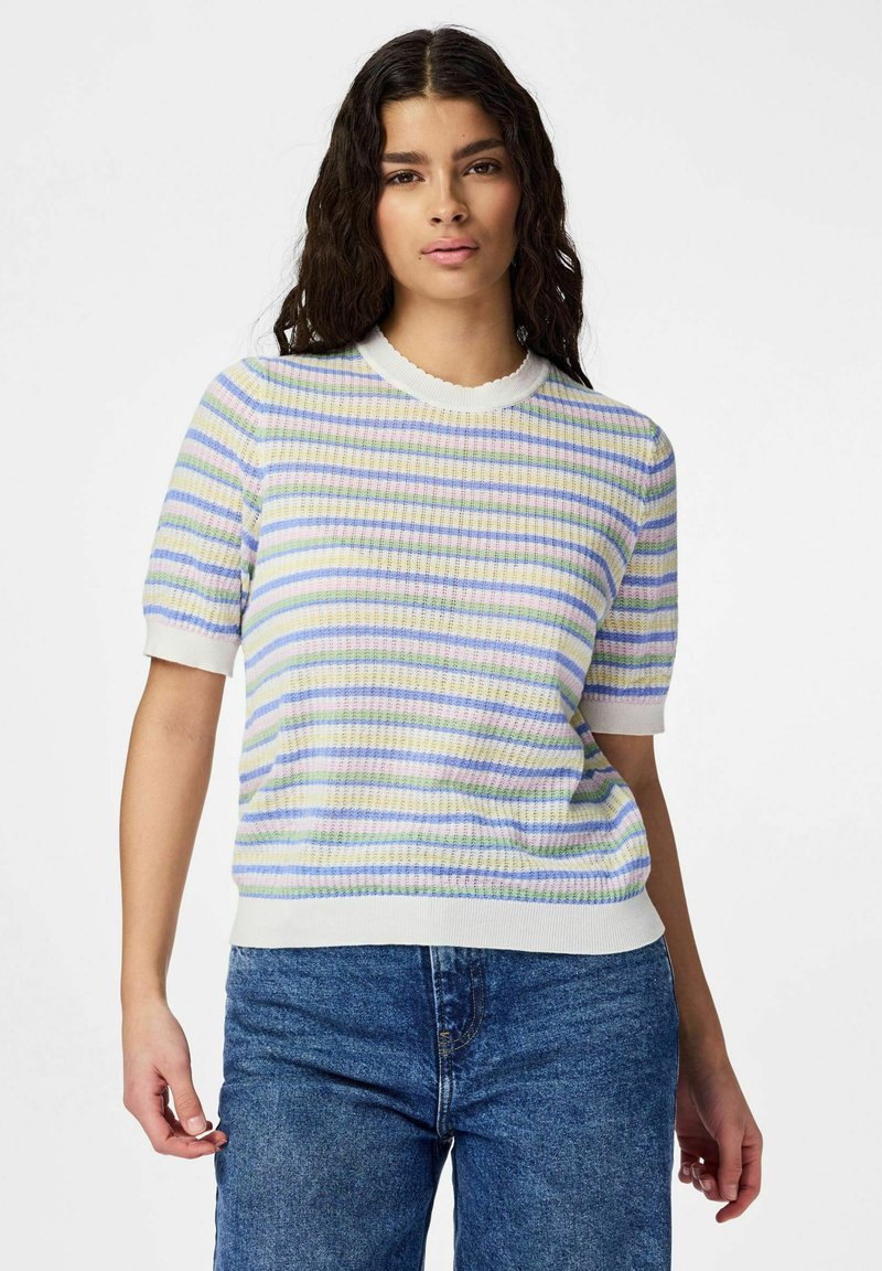 Femme aux cheveux bouclés foncés, portant un haut tricoté à manches courtes à rayures pastel et un jean bleu, debout devant un fond blanc uni.