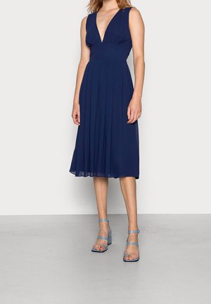 Robe sans manches bleu marine avec un décolleté en V plongeant et une jupe plissée, assortie à des sandales à talons bloc à brides bleu clair.