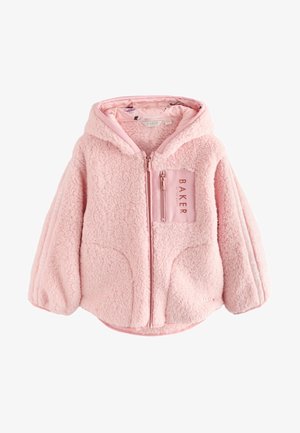 Smilende pige iført en oversized blød lyserød pelsagtig hættejakke over hvid skjorte og sorte leggings, stående mod en ensfarvet lys baggrund.