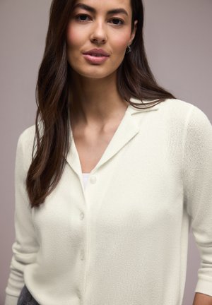 Femme aux longs cheveux bruns portant un cardigan blanc boutonné et un haut blanc sur un fond neutre uni.