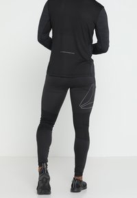 Mężczyzna w czarnych legginsach sportowych z geometrycznym wzorem na udzie i czarnej bluzie z długim rękawem, stojący tyłem na białym tle.