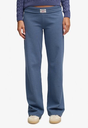Personne portant un pantalon large à fines rayures bleues et noires avec une étiquette Superdry à la taille et des baskets beiges.
