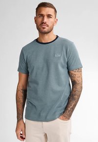 Tricou texturat, albastru, cu mâneci scurte și decolteu rotund bleumarin, cu o mică emblemă pe piept; purtat de un model cu tatuaje.