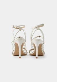 Sandales à talons hauts blanches avec un design à brides croisées, des côtés ouverts et une finition brillante. Elles présentent un talon effilé et une bride de cheville avec boucle.