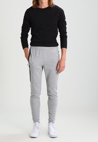 Schwarzes Langarmshirt kombiniert mit grauen Slim-Fit-Jogginghosen mit Reißverschlusstaschen, flachem Bund und weißen Sneakern.