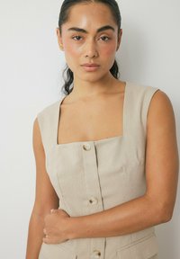 Beige linnedress med firkantet udskæring og uden ærmer. Har frontknaplukning og sidelommer, samt en tekstureret overflade.