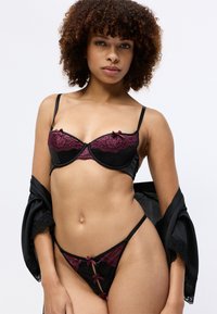 Zwarte satijnen lingerie set met bordeauxrode kanten rand, met strikjes, beugelbh en bijpassende string. Zachte textuur en fijne details.