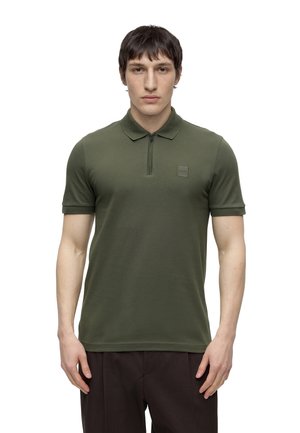 Ung mand iført en olivengrøn kortærmet polo med lynlås og lille brystlogo, stående mod en hvid baggrund.