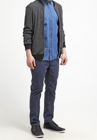 Conjunto de hombre que incluye una chaqueta bomber texturizada en gris, una camisa vaquera de botones, chinos en azul marino y zapatillas negras con acentos grises.