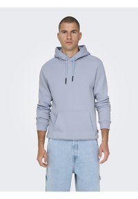 Sudadera ligera gris claro hecha de una tela suave, que presenta un bolsillo frontal, capucha con cordón y puños de canalé. Combinada con pantalones cortos de mezclilla azul claro.