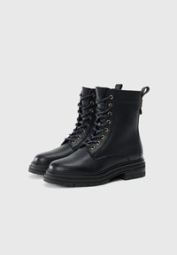 Botas de combate de cuero negro con cordones, suelas de goma gruesas y ojales dorados, mostradas en ángulo lado a lado sobre un fondo blanco.
